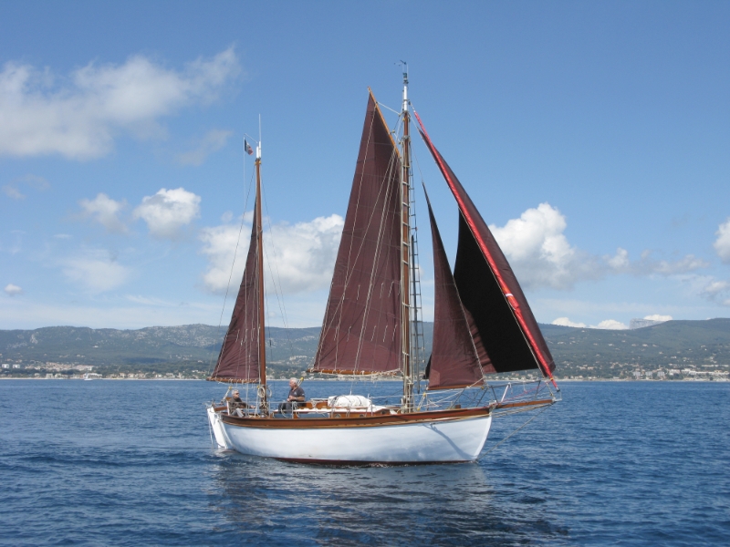 Un voilier en mer avec des voiles marron, naviguant sur des eaux calmes sous un ciel ensoleillé, avec des montagnes en arrière-plan.