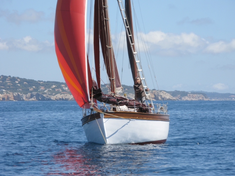 Un voilier avec une grande voile colorée naviguant sur l'eau calme, avec un marin à bord et un paysage côtier en arrière-plan.
