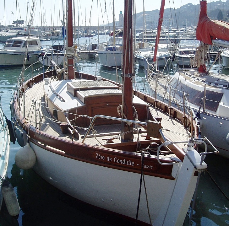 Un voilier amarré dans un port, nommé 'Zéro de Conduite', avec une coque blanche et un pont en bois, entouré d'autres bateaux.