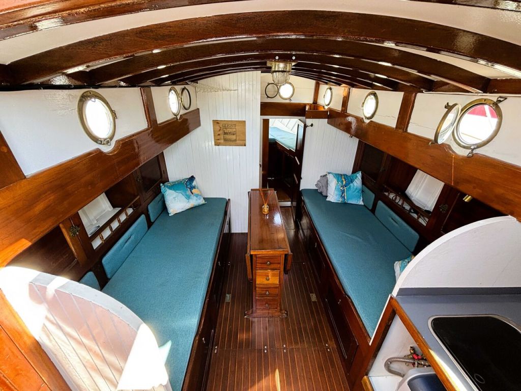 Intérieur d'un bateau avec deux banquettes longues. Décor en bois, fenêtres circulaires et table au centre.