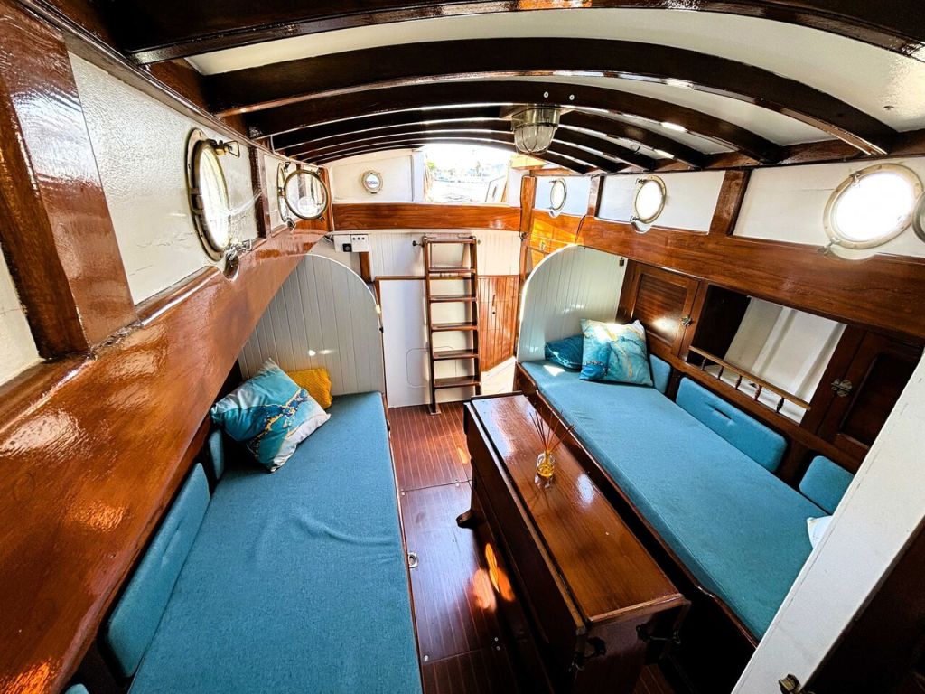 Intérieur d'un voilier avec deux banquettes bleues, une table en bois, des hublots ronds et des étagères en bois.