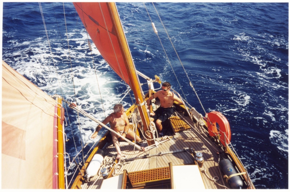 Deux marins sur le pont d'un voilier avec des voiles rouges, naviguant sur une mer bleue.