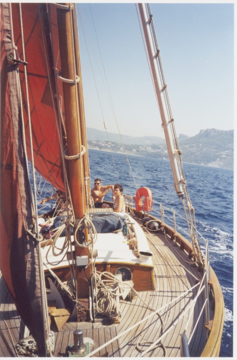 Deux personnes sur le pont d'un voilier naviguant en mer, avec des montagnes en arrière-plan et une voile rouge au premier plan.