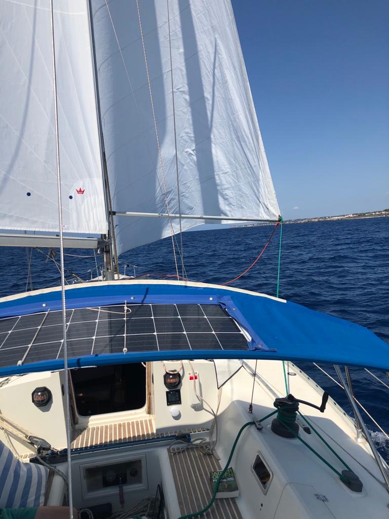 Vue sur un voilier avec des voiles blanches, le toit du cockpit équipé de panneaux solaires, naviguant sur une mer bleue claire.