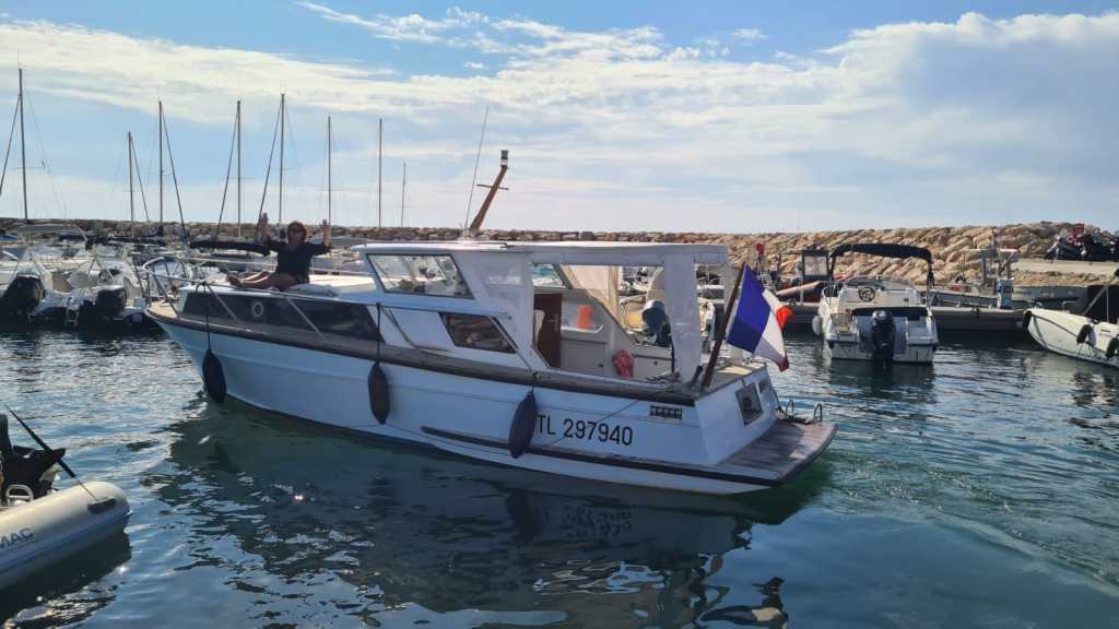 Un bateau à moteur blanc se déplace dans un port, entouré de plusieurs autres petites embarcations. Une personne est assise à l'avant, levant les bras en signe de joie, tandis qu'un drapeau français est visible à l'arrière du bateau.