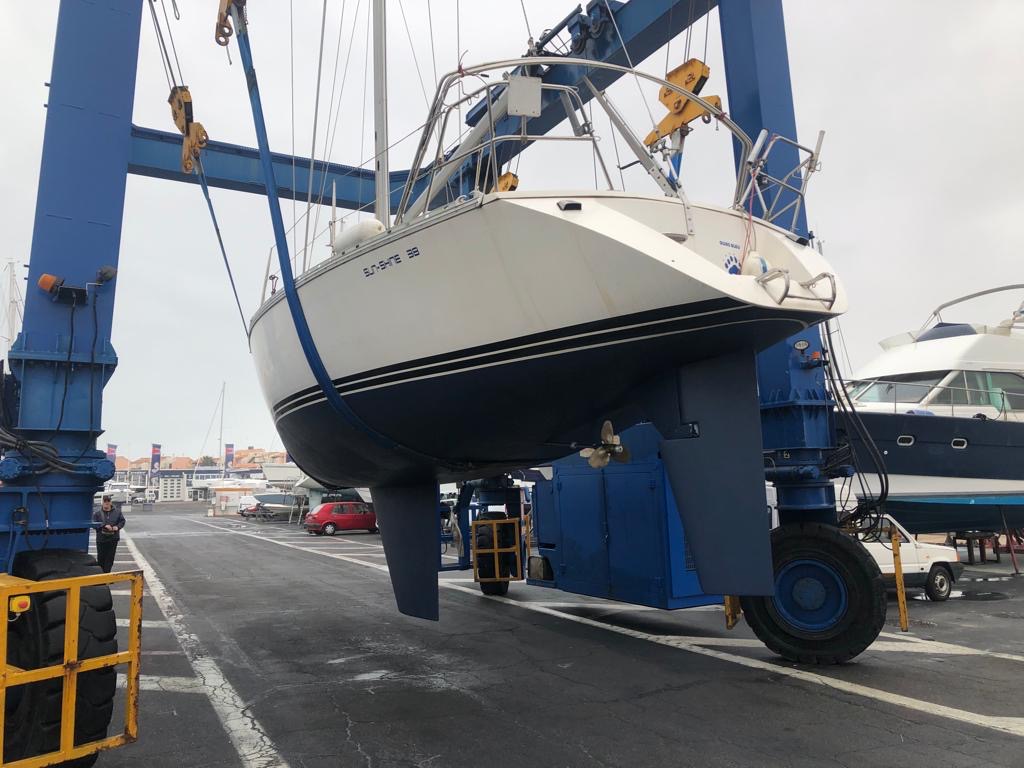 Un bateau suspendu en l'air par une grue au port, avec d'autres bateaux et des voitures en arrière-plan.