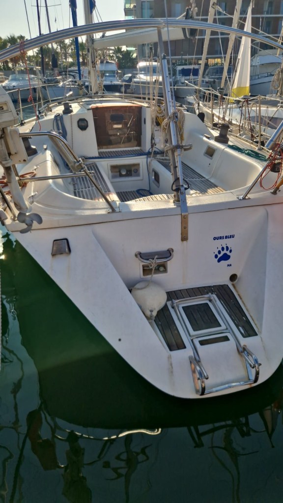 Vue arrière d'un voilier amarré dans un port, avec un cockpit et un logo bleu en forme de patte sur le côté.