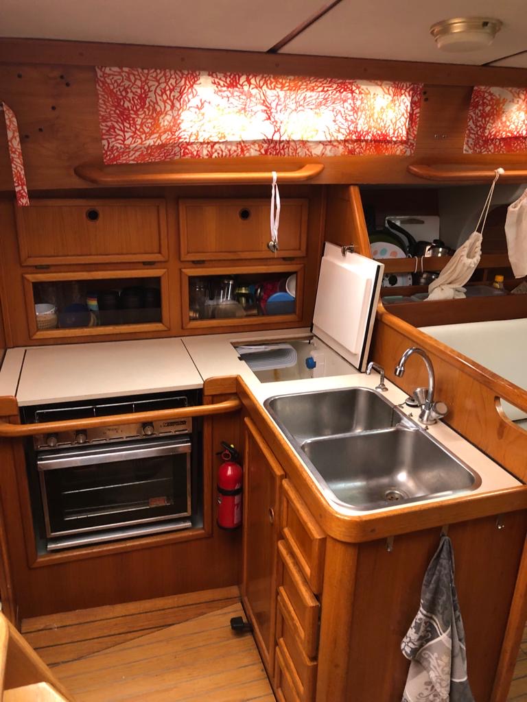 Cuisine d'un bateau en bois avec évier, four et rangements, éclairée par une fenêtre ornée de tissu.