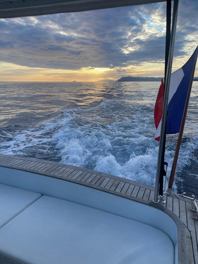 Vue d'un bateau en mer avec un ciel nuageux et un coucher de soleil à l'horizon, drapeau français visible.