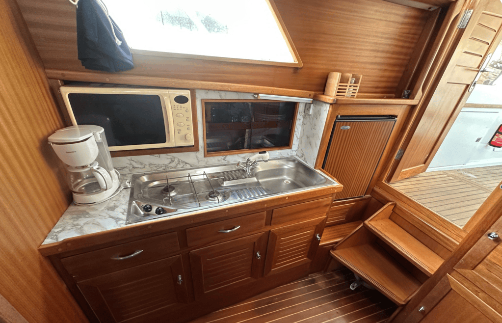 Cuisine intérieure sur un bateau, avec un évier, une cuisinière à gaz, un micro-ondes, une cafetière et un petit réfrigérateur en bois.