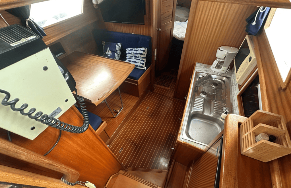 Vue intérieure d'un bateau, montrant la cuisine avec un évier, une cafetière et une table en bois. Les murs en bois et les espaces de rangement sont visibles.