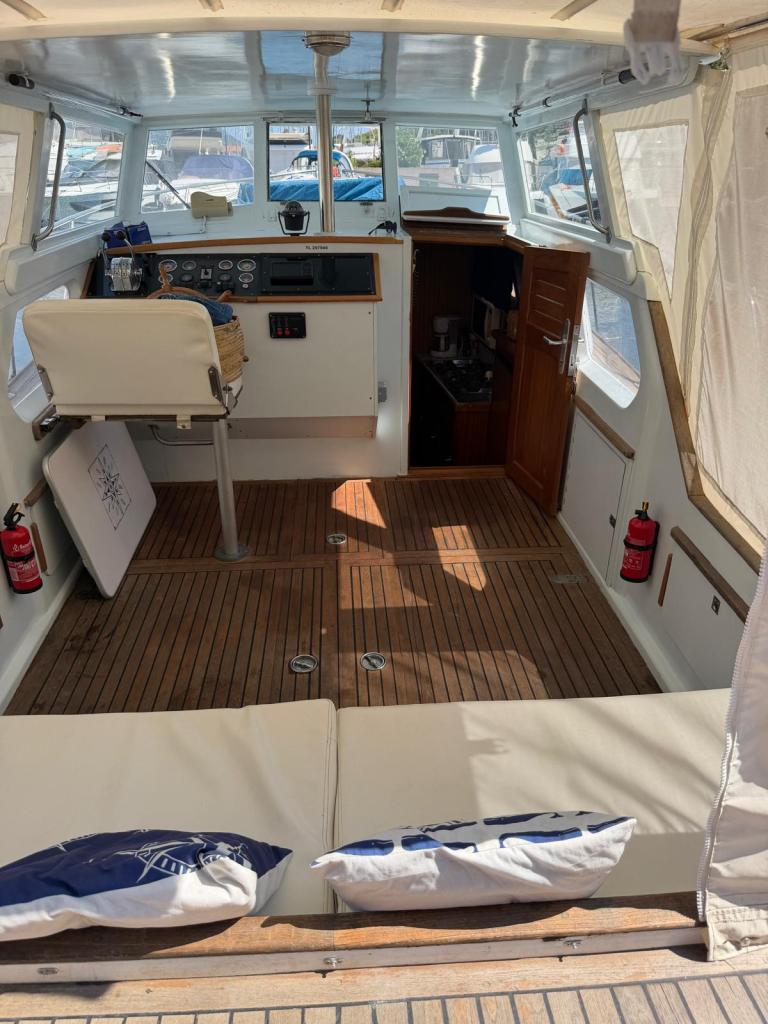 Intérieur d'un bateau avec un tableau de bord, un fauteuil de pilotage et des coussins sur le pont en bois.
