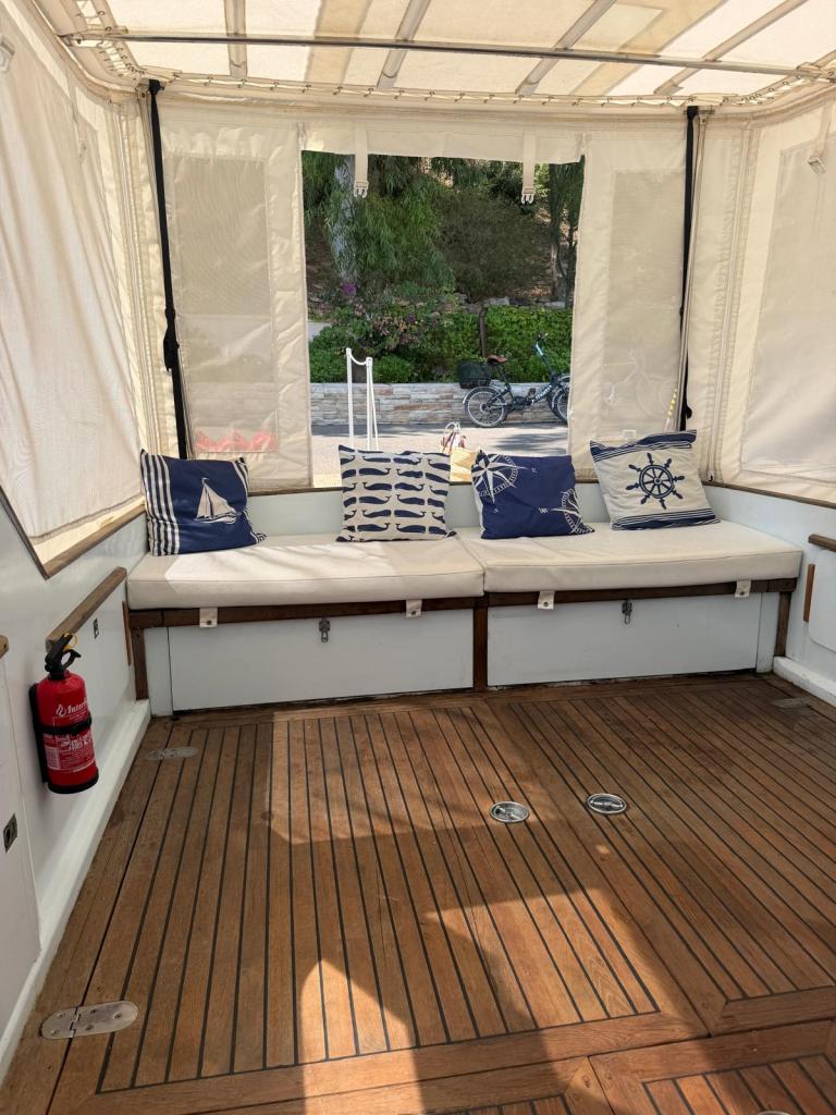 Intérieur d'un espace confortable sur un bateau, avec des coussins marins et un sol en bois, offrant une vue sur l'extérieur.